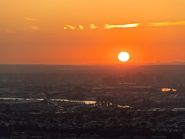 New York City, United States – October 2024: Panoramic sunset view over New York City, showing the sun descending beyond the horizon and casting a warm orange glow over the urban landscape. The image captures bridges, waterways, and distant neighborhoods under the fading light of day. A commercial airplane is visible in the sky, emphasizing the dynamic character of the city and its connection to global travel.