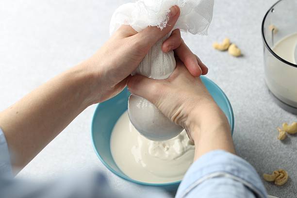 istock Woman straining cashew milk into bowl at light table with nuts, closeup 2243100013