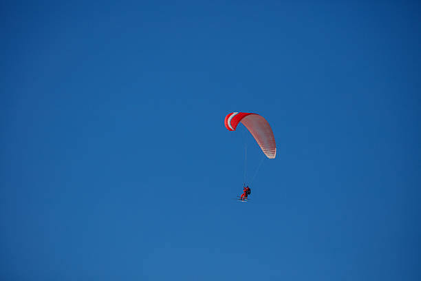 Paraglider skiers, instructor and novice glide silently across a clear cobalt sky, suspended in crisp mountain air on red wing