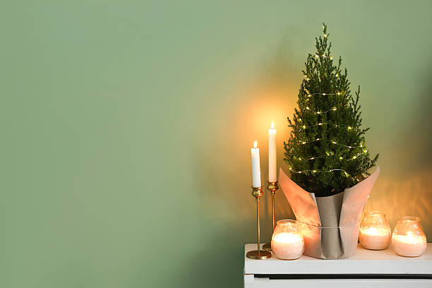 istock Small spruce tree decorated with Christmas lights and burning candles on white radiator cabinet near green wall, space for text 2243111092