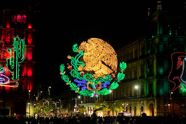 Nighttime scene in Mexico City’s Zocalo showing the Mexican national emblem made of colorful lights glowing over historic buildings while crowds enjoy the Independence Day festivities, national pride