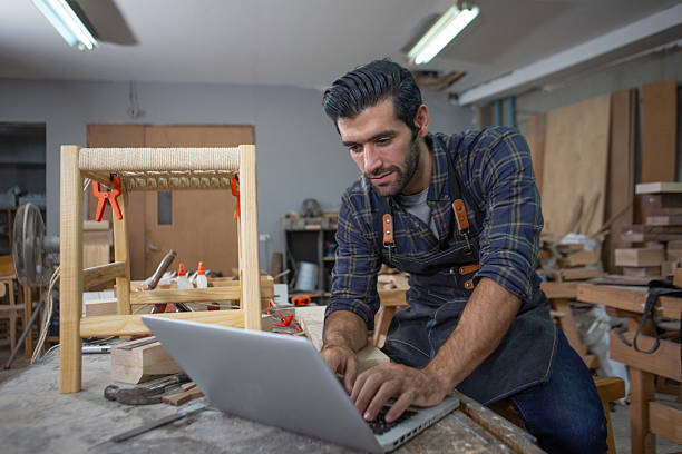 istock young carpenter caucasian man with laptop make drawing on wood to design at workspace. craftsman profession in wood factory 2243118111