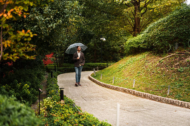 Young man holding a transparent umbrella while walking along a curved path surrounded by trees and greenery. He wears jeans and a brown jacket, enjoying a quiet rainy day in a park environment.