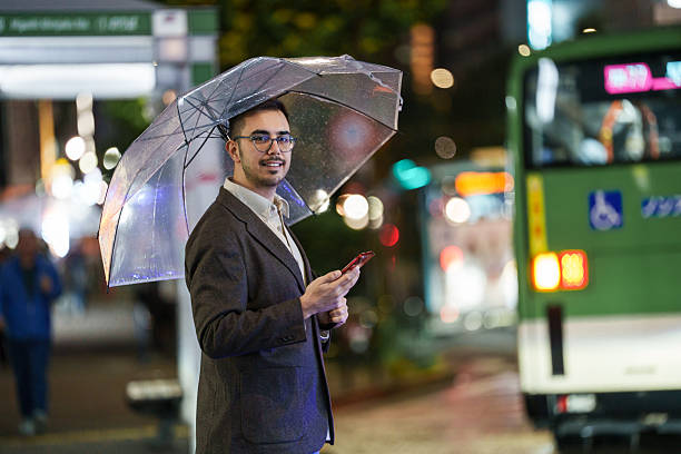 Young man holding a transparent umbrella and checking his smartphone while standing near a city bus on a rainy night. Bright streetlights and reflections from wet pavement create an urban evening atmosphere.