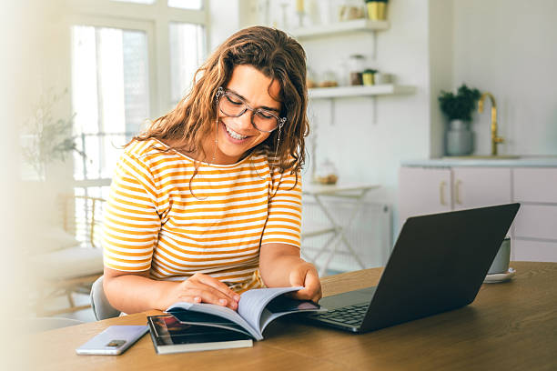 istock Smiling woman reading book at kitchen table with laptop and coffee in bright modern interior 2243131447