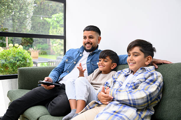 Cheerful British Pakistani father and two boys relaxing at home, sitting on the sofa, watching television, fun, enjoyment, leisure
