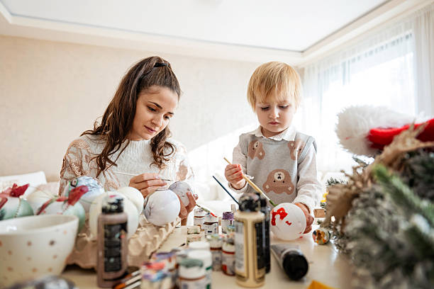Mother and her little son painting Christmas tree decorations at home stock photo