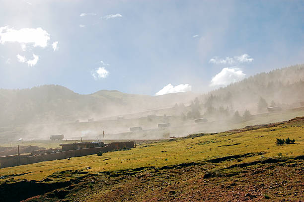 A misty rural landscape featuring green fields and distant mountains under a clear sky with scattered clouds.