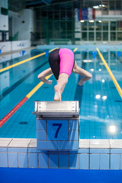 Female swimmer in professional swimsuit diving into the water from a starting block during a swimming competition at an indoor pool. Sports action moment symbolizing determination, focus, and athletic performance in competitive swimming.