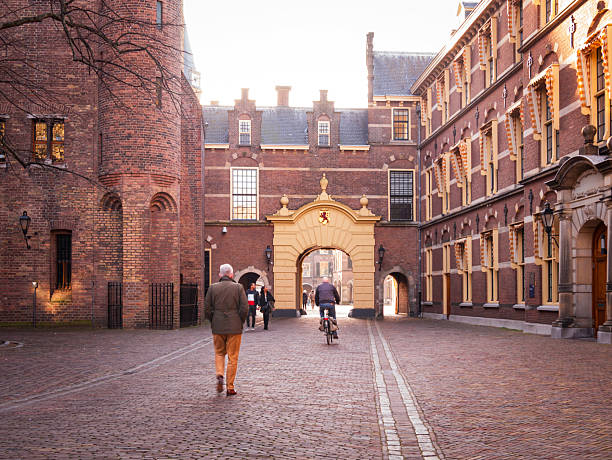 The Hague, Netherlands – February 17, 2019: The historic Binnenhof complex with brick buildings, arched gateway, and people walking and cycling on the cobblestone courtyard.