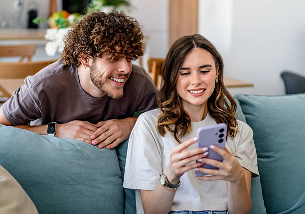 istock Young couple smiling sharing smartphone on sofa at home 2243203080