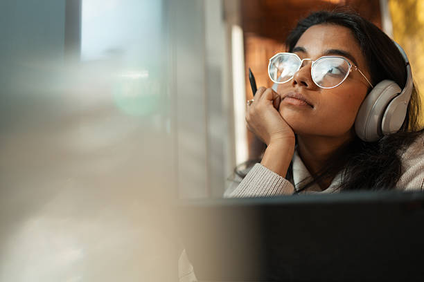 Young woman in glasses and headphones, holding a pen and looking away thoughtfully, perhaps studying or brainstorming