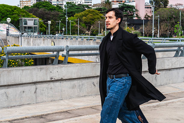 Young man wearing a long coat and jeans walking purposefully on a concrete bridge in a city environment