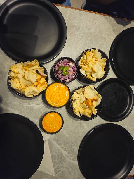 An appetizing overhead view of a table set with bowls of crispy papad (or khichiya papad), a beloved Gujarati snack. The papads are served alongside a small bowl of finely chopped red onion and green chili salad and bright orange-yellow curd dip . Several empty black plates are arranged, ready for service
