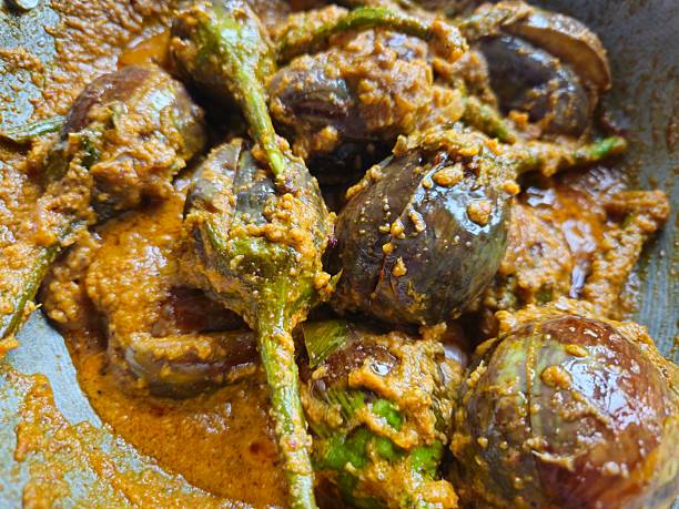 A close-up view of a traditional Indian curry in the making. Baby brinjals (eggplants) are simmering in a rich, thick, orange-brown gravy made from peanuts, coconut, and spices