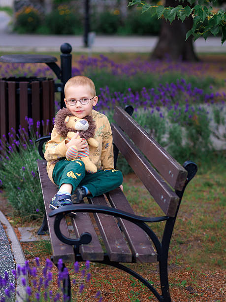 Serious boy with toy lion sitting alone on bench surrounded by lavender flowers. Concept of childhood emotion, reflection, and calm atmosphere.