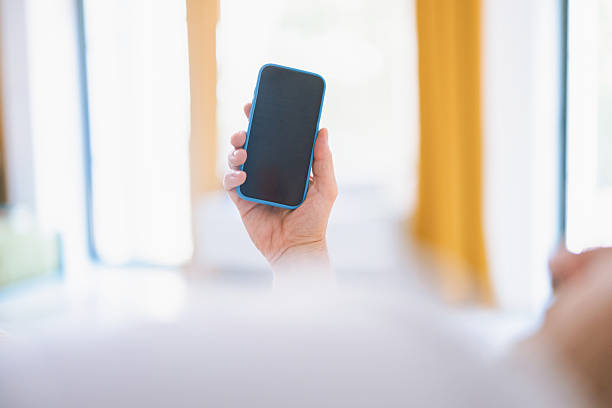 A close-up of a hand gripping a blue smartphone with a blank screen, raised in a bright, softly blurred indoor setting. Ideal for tech, mobile, and communication visuals.