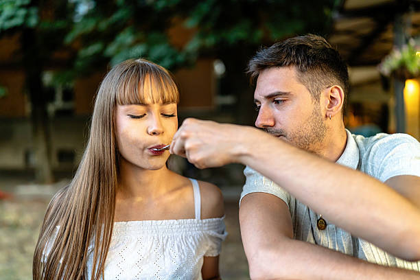 A man and woman sit together outdoors, sharing food in a loving gesture. The interaction captures intimacy and the joy of spending quality time together against a tranquil backdrop, symbolizing romance and connection.