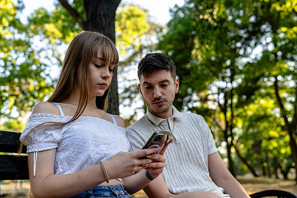 istock Young couple interacting with a smartphone on a bench in a sunny park 2243293375