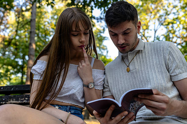 A young couple sits closely on a park bench, immersed in reading a book together. The serene outdoor setting of tall trees lends an idyllic atmosphere. Their gestures and expressions show a focused and engaged connection, enhancing the intimate and thoughtful scene. This moment captures shared interest, intellectual curiosity, and companionship in a peaceful environment.