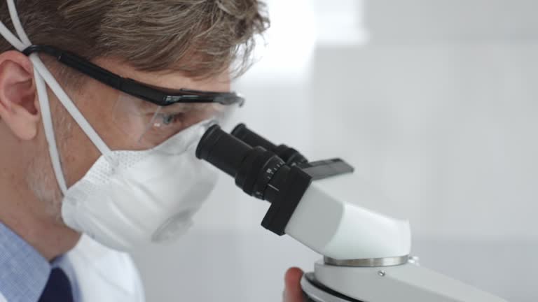 Male scientific researcher wearing protective gear examining sample through microscope, performing detailed laboratory investigation. Medicine and science concept