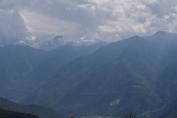 Snow-capped peaks of the Italian Alps rise prominently above lush, forested slopes. The snow contrasts sharply with the dark green of the trees. Fluffy clouds float in the bright blue sky above, partially obscuring some peaks and casting soft shadows on the landscape. The rugged texture of the mountains adds depth and drama to the scene. The interplay of light and shadow enhances the natural beauty of this alpine vista.