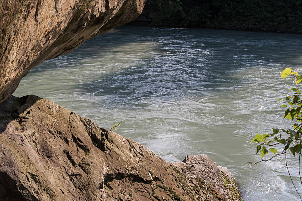 A rocky cliff features prominently on the left, with rugged textures and sunlit surfaces. Beside it, a river flows, displaying swirling greenish-blue water. The calm atmosphere is accentuated by sunlight filtering through foliage, casting shadows on the rocks. Vegetation appears in the background, contributing to the serene natural setting. This scene reflects a peaceful interaction between geological formations and flowing water.