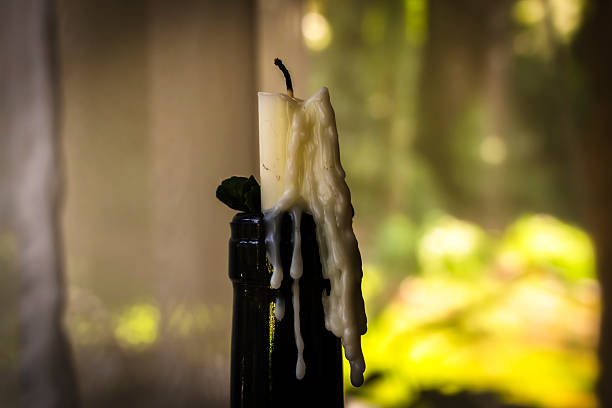 An extinguished white candle with melted wax dripping down a dark glass bottle. The scene is set against a brightly lit window with a soft-focus background, evoking a sense of nostalgia and passed