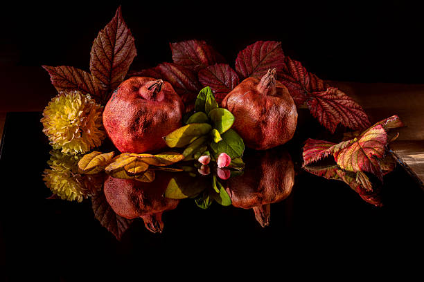 Two pomegranates with autumn leaves, flower, and small buds arranged creatively on shiny black surface. Harvest season concept. Rosh Hashanah. In low key.