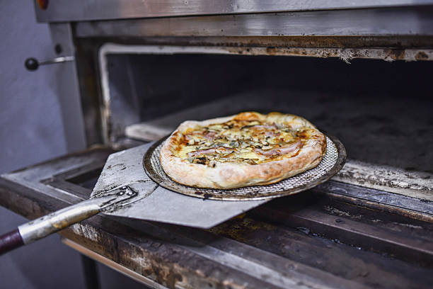 A busy kitchen scene shows a chef sliding a freshly baked pizza into an industrial oven using a metal pizza peel. Warm, delicious, and ready to serve in a professional setting.