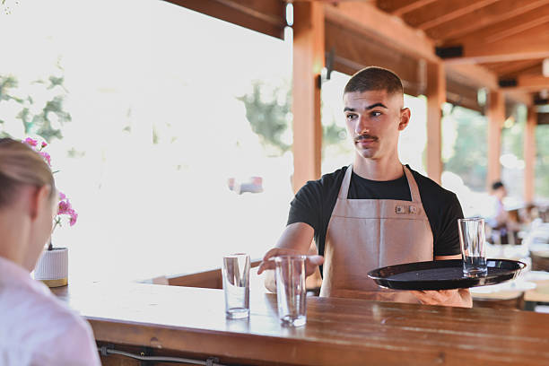 A female bartender in a white shirt and waiter are preparing and hands drinking glasses for a customer at a wooden bar. Refrigerated beverages line the back wall, capturing a casual hospitality moment.