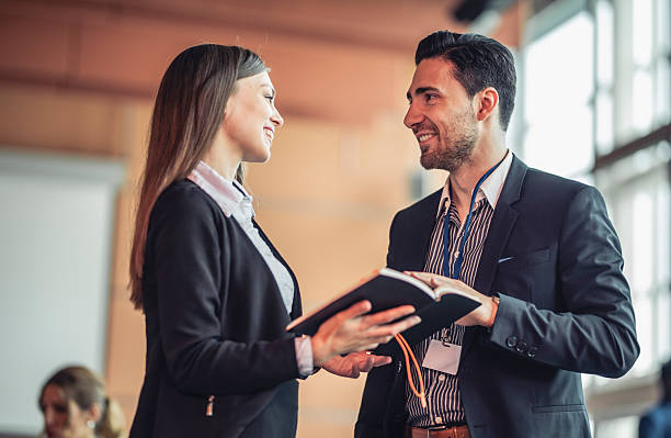 Two Caucasian business executives in formal attire engage in a lively discussion at a conference. They are smiling and holding an open notebook, indicating an exchange of ideas. A projector screen is visible in the background.