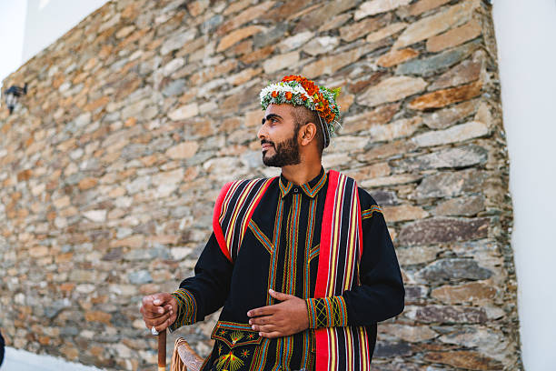 Middle Eastern man standing against a rustic stone wall, wearing traditional colorful attire and a floral headpiece. He is holding a wooden cane, looking off to the side with a thoughtful expression. The setting suggests a historical or cultural location.