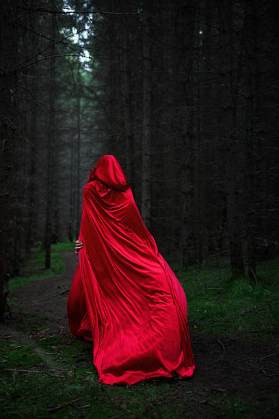 istock figure clad in a flowing red cloak wanders through a dark forest. Tall trees surround the path, creating an eerie yet captivating atmosphere as twilight descends 2243317581