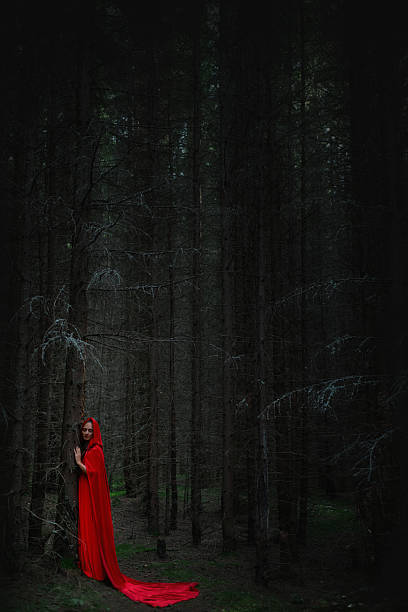 istock model dressed in a flowing red gown stands against a tree in a dimly lit forest. surrounding trees cast shadows, adding a mystical vibe to the scene at dusk 2243317642