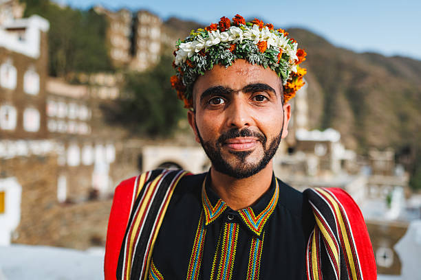 Portrait of a Middle Eastern man wearing a colorful floral headpiece and traditional Asiri attire. He is standing outdoors at the Rijal Almaa heritage site, with historic buildings visible in the background. The scene captures the cultural essence of the location.