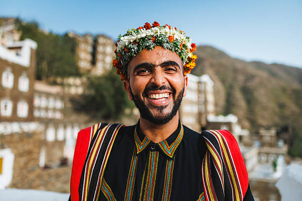 Smiling Saudi tour guide wearing colorful traditional attire standing outdoors at Rijal Almaa heritage site with historic buildings in background.