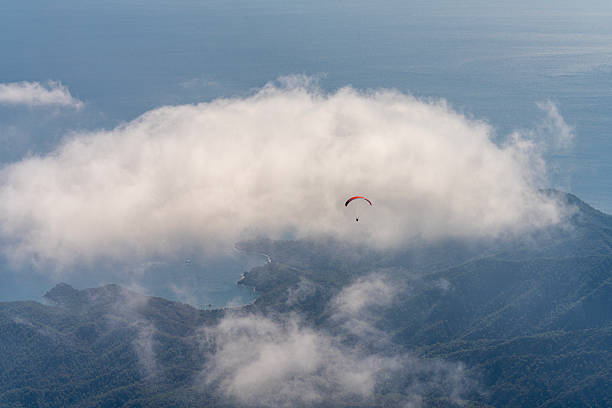 A high-angle, atmospheric shot of a single paraglider with an orange canopy soaring over a dense, foggy coastal landscape