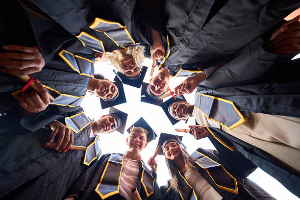 Diverse group of young adult students looking down, celebrating graduation while holding diplomas and standing in a huddle