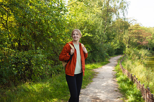 Happy young woman with a backpack wearing an orange jacket walking along a forest path on a sunny day. Concept of hiking, outdoor adventure, travel, freedom, and healthy lifestyle.
