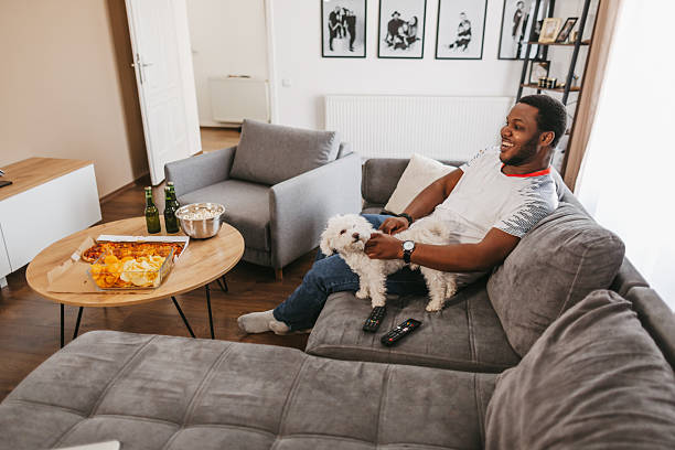 istock Young man with beer and snacks sitting on sofa and petting his little dog while watching match on TV at home 2243352204