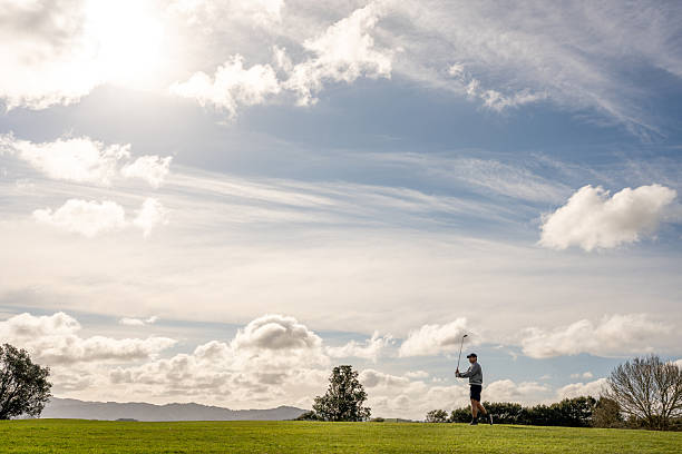 A male golfer stands on the fairway holding a club, with the sun shining through scattered clouds