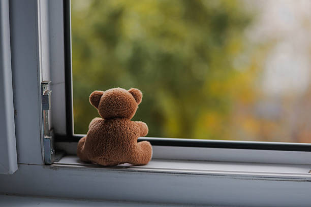 istock Sad brown teddy bear alone sitting on open window sill and looking out of home. 2243375151