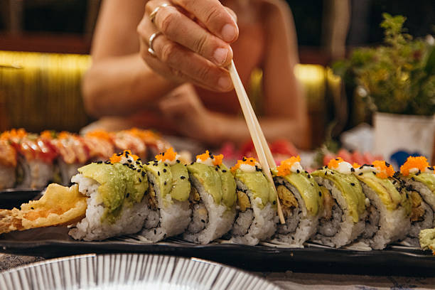 Hand using chopsticks to pick a sushi roll topped with avocado and fish roe. Fresh Japanese sushi on black plate with various rolls in restaurant setting. Asian cuisine dining experience.
