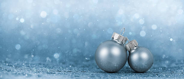 Elegant close-up of two silver Christmas ornaments placed on a frosty blue glitter surface with soft bokeh lights in the background. The cool tones and minimal composition evoke a peaceful winter mood, ideal for Christmas and New Year greeting cards, seasonal advertisements, or elegant winter-themed designs with copy space.