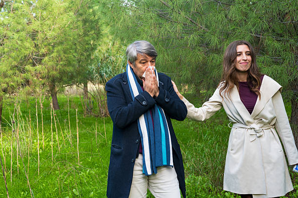 Outdoor scene with a caucasian middle-aged man sneezing into a tissue while a younger woman gently supports him, illustrating early cold or flu symptoms in a natural park environment.