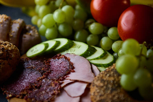 A cinematic still life photo showing ham, salami, tomatoes, grapes, bananas, and cinnamon buns arranged on a table with moody light emphasizing color and texture.