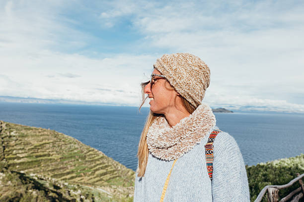 Smiling woman in knit hat and scarf standing near ocean with hills in background. Happy young adult enjoying cold weather and nature. Outdoor lifestyle and winter fashion concept.