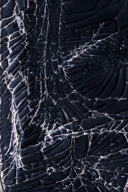 Macro close-up of shattered safety glass on a deep black background, revealing sharp splinters and spiderweb-like fracture lines radiating from an impact point. High-contrast abstract texture with natural copy space, ideal as a dramatic background or design element.