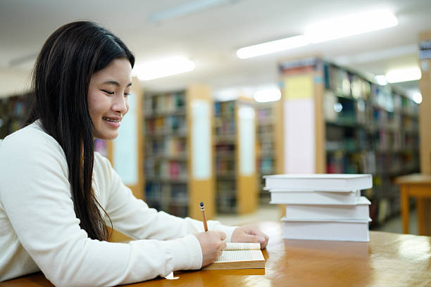 istock Asian young Student doing homework in library of university or college with various book and stationary over the book shelf background, Back to school 2243407110